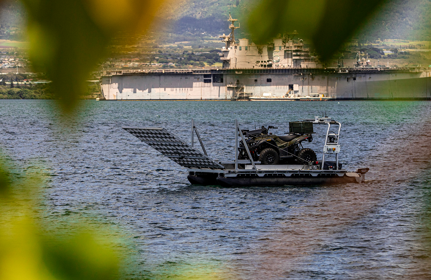 an atv being carried on an unmanned autonomous boat with a battleship in the background